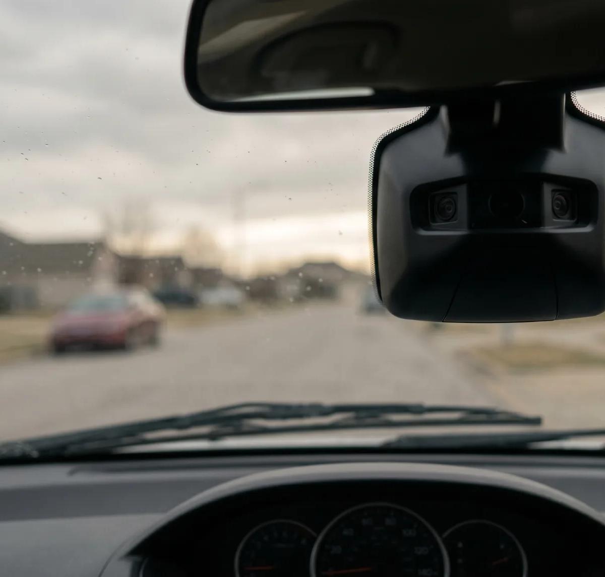 Driver's point-of-view through the windshield of a mid-size SUV showing the dual-lens front-view camera module mounted below the rearview mirror, with a quiet suburban street and a distant red car visible ahead under an overcast sky.