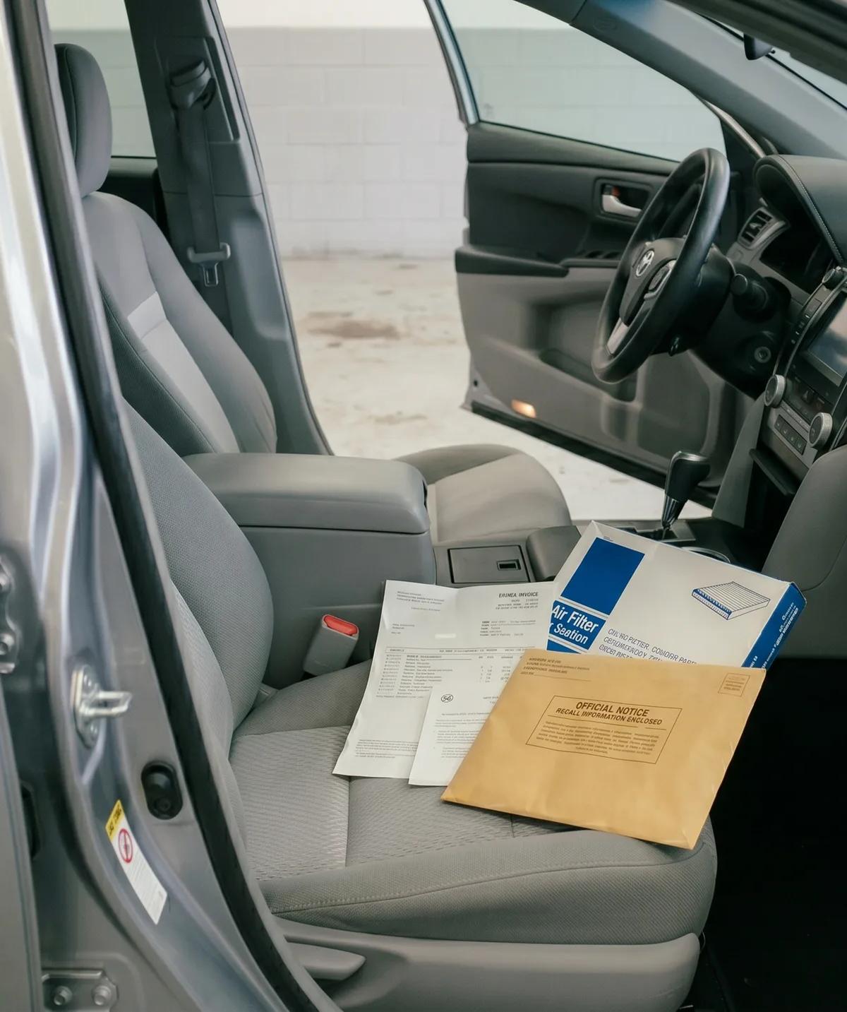 Open passenger door of a Toyota Camry sedan revealing a service invoice, a cabin air filter in its blue box, and a manila envelope marked "Official Notice — Recall Information Enclosed" resting on the gray cloth passenger seat, with the Camry's dashboard and steering wheel visible in the background.